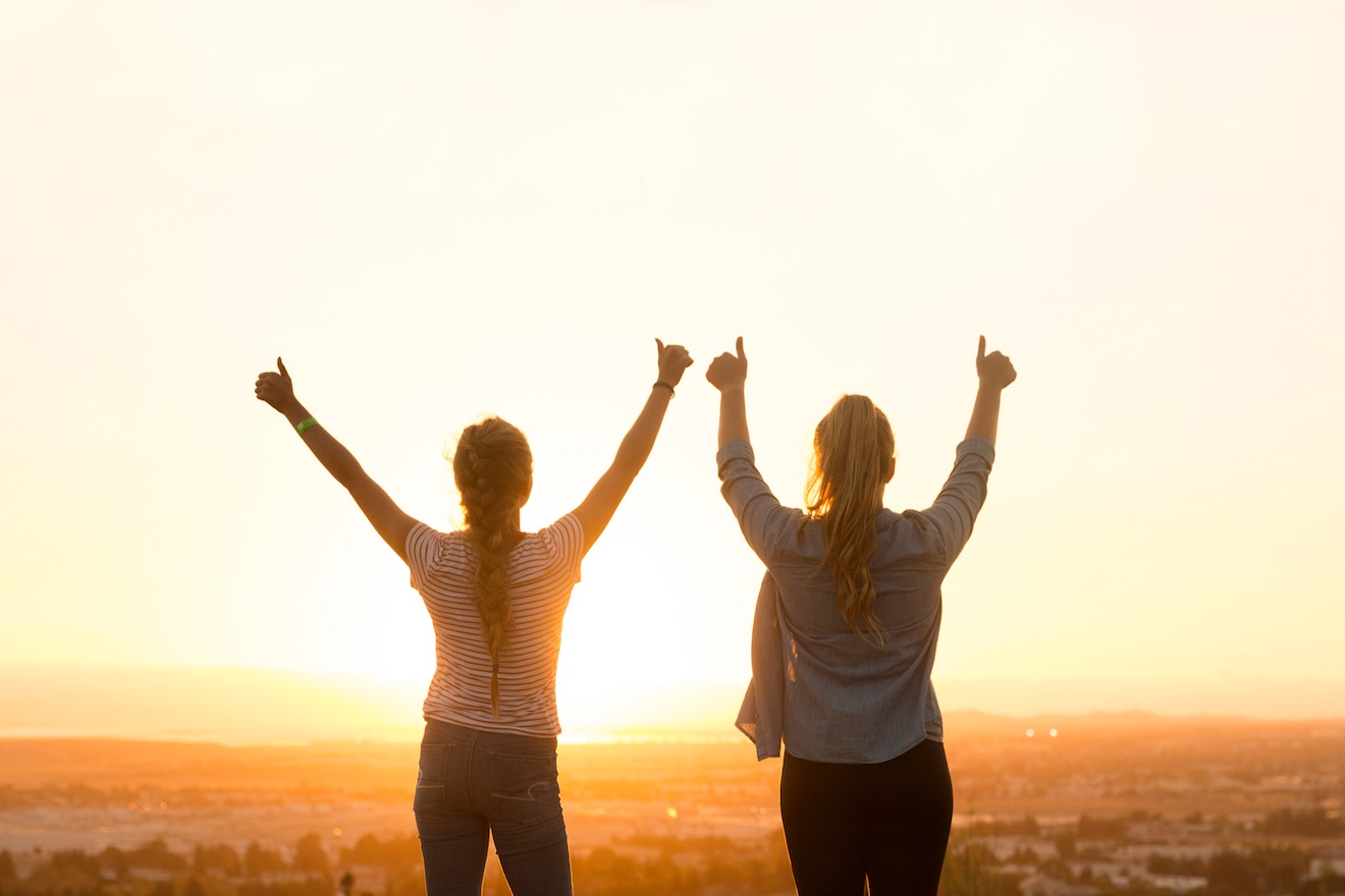 Two women with arms up in front of sunset